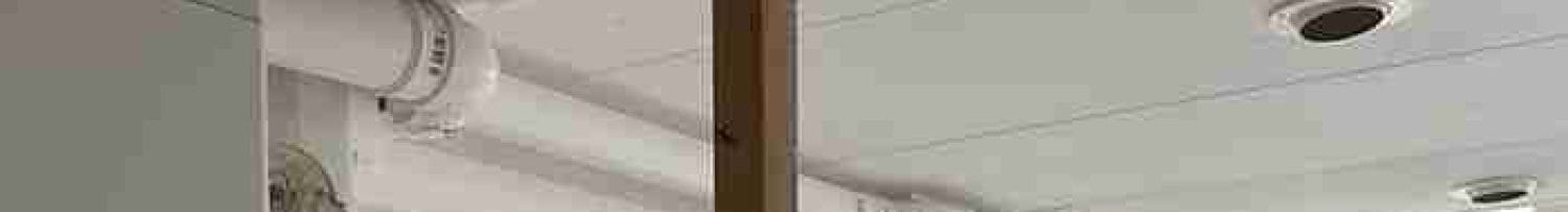 Plafond met witte leidingen, ventilatieopeningen en houten balk